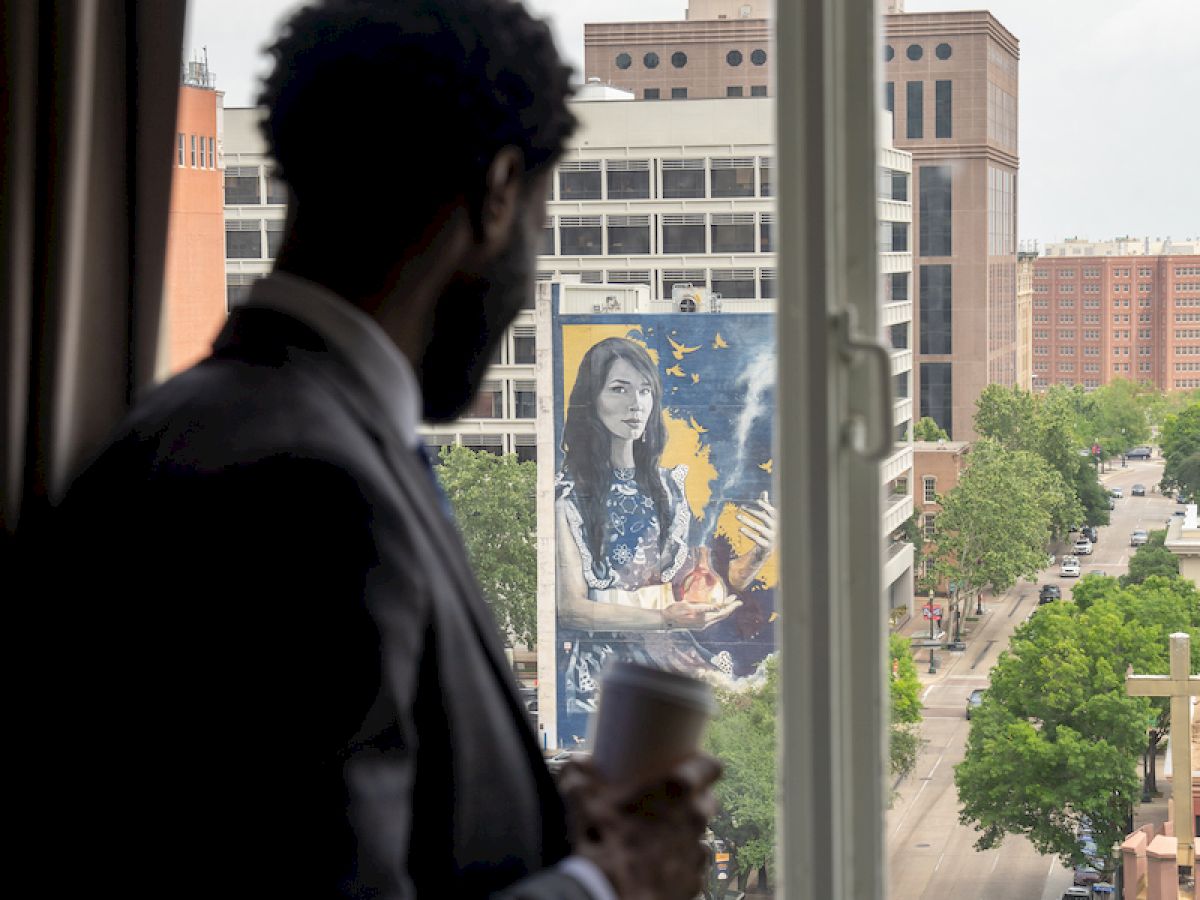 A person stands by a window holding a cup, looking at a cityscape with buildings and a mural in the background.