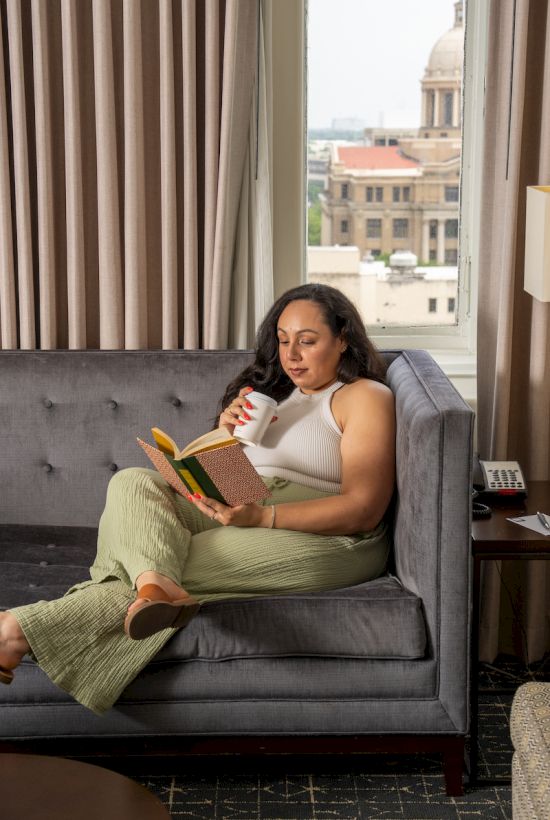 A person is sitting on a gray couch, reading a book and drinking from a mug, with a view of buildings through a large window.