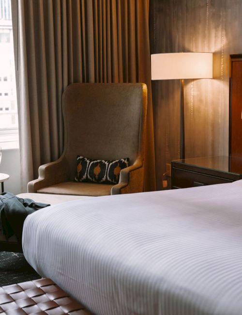 The image shows a hotel room with a large bed, an armchair, a small table with a glass, and a window with curtains.