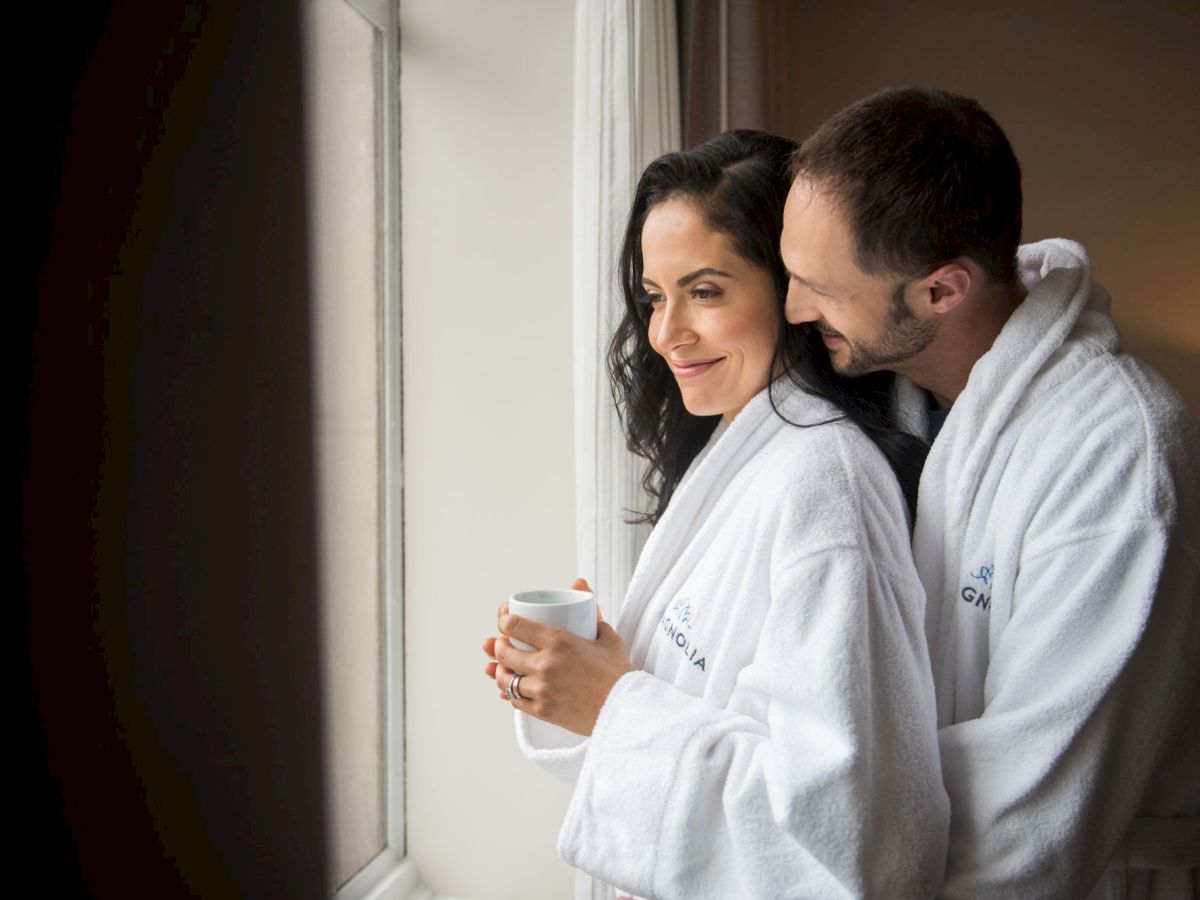A couple wearing white robes, one holding a mug, stands by a window looking outside.