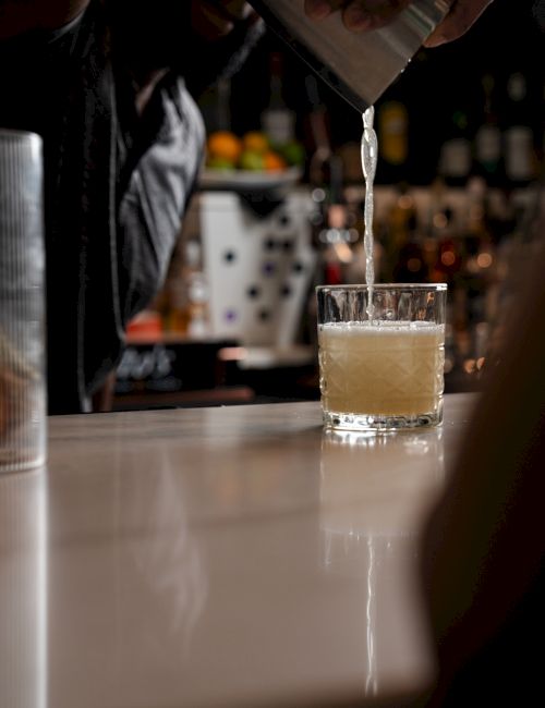 A person pours a drink from a shaker into a glass on a bar counter, with a blurred background of bottles and light.