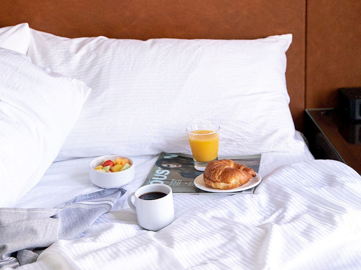 A bed with white sheets has a breakfast setup, including a croissant, coffee, juice, and fruit salad. A horse photo is above the bed.