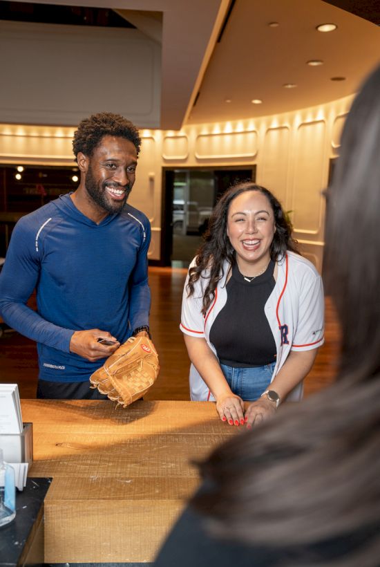 Two people are smiling at a counter, with one holding a baseball glove. Another person is partially visible in the foreground.