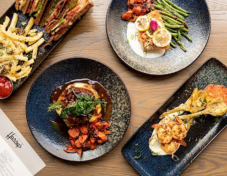 The image shows four dishes on black plates, including fries, seafood, and meat, with a menu on a wooden table.