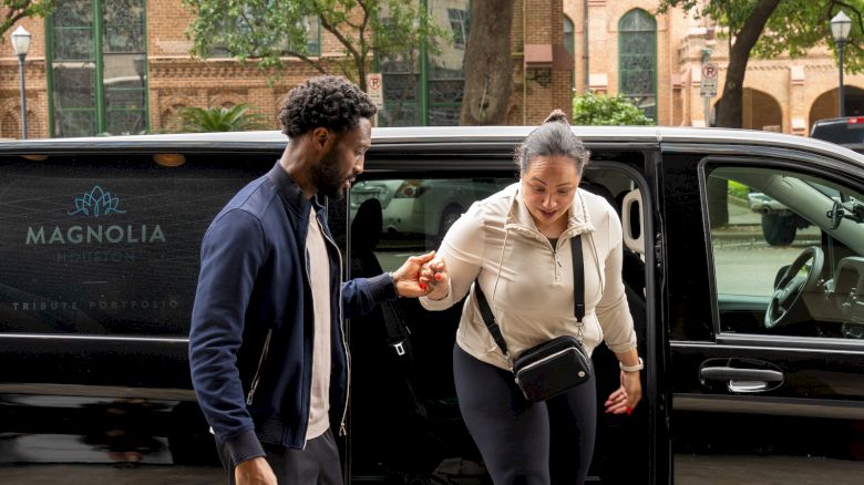 A person helps another out of a black van in front of a brick building with arched windows and trees lining the street.
