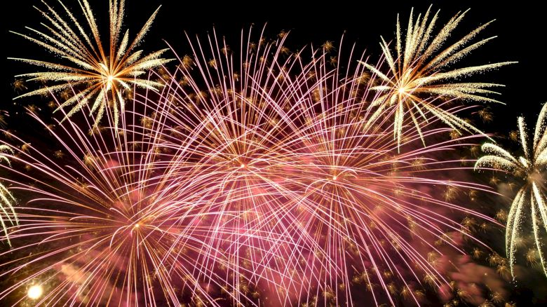 The image displays a vibrant fireworks display against a dark night sky, with bursts of pink and gold illuminating the scene.