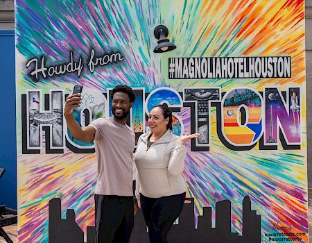 Two people pose in front of a colorful mural reading 