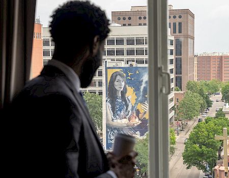 A person stands by a window holding a cup, gazing at a city view with a large mural on a building across the street.