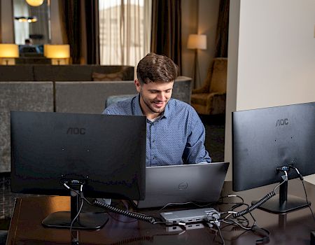 A person is working at a desk with a laptop and two monitors, in a room with curtains and lamps in the background.