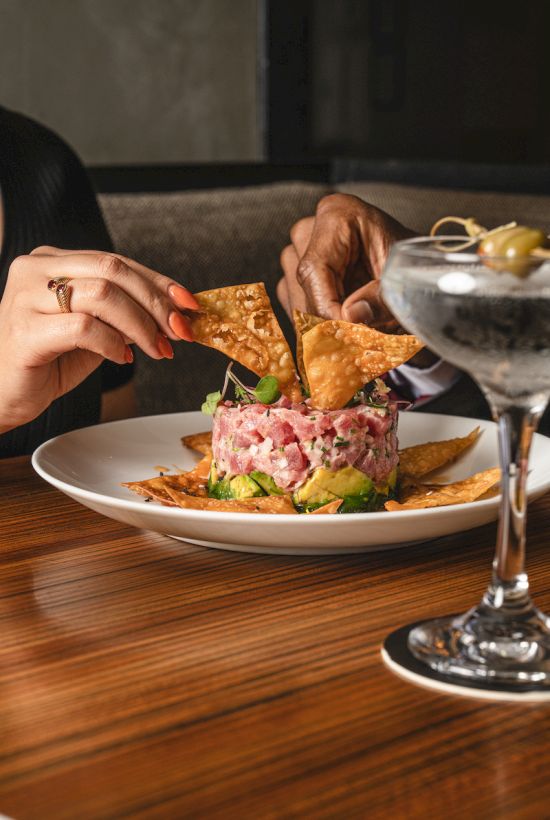 Two people share a dish with tortilla chips and a mixed topping beside a glass with an olive garnish on a wooden table.