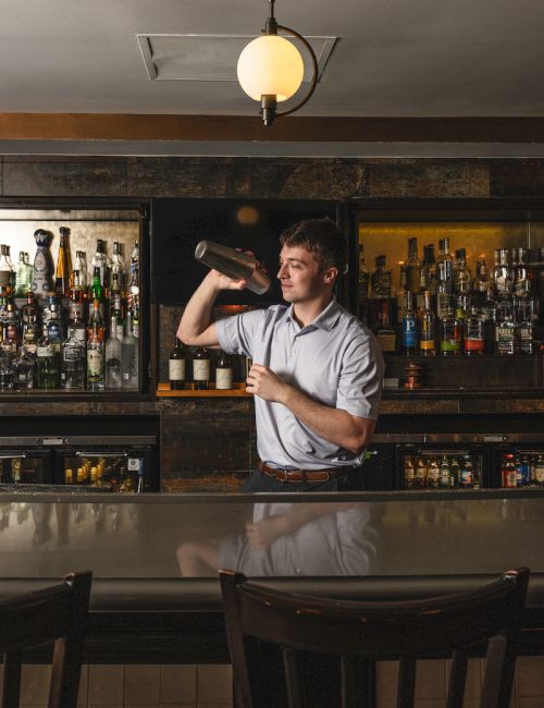 A bartender is shaking a cocktail shaker behind a bar, surrounded by shelves of bottles, under a ceiling light in a cozy setting.
