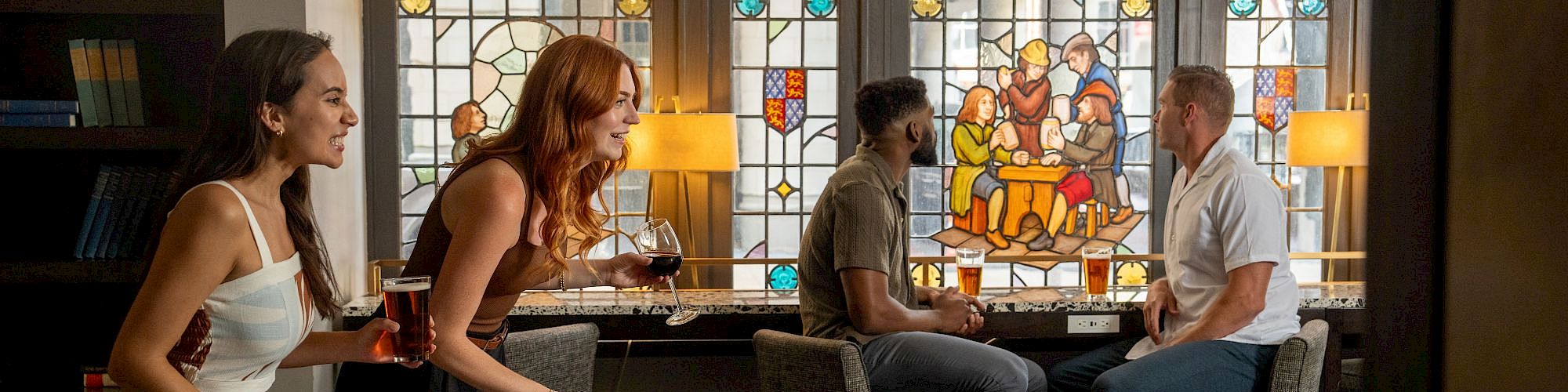 People are socializing in a room with stained glass windows and a game table. Two are playing, while others chat in the background.