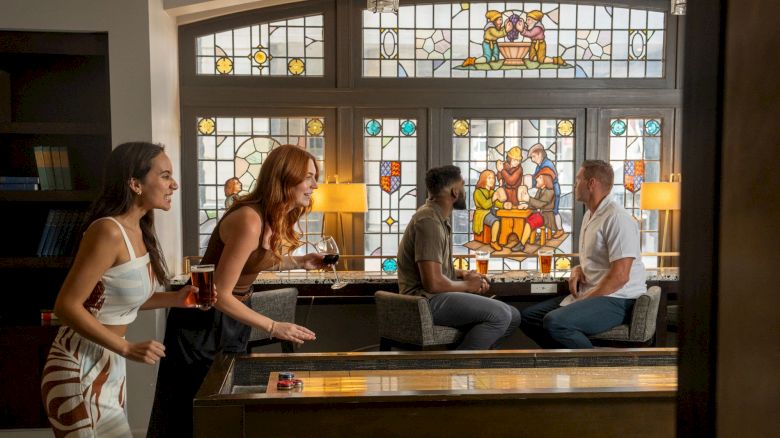 People are enjoying drinks and playing shuffleboard in a room with colorful stained glass windows depicting a biblical scene.