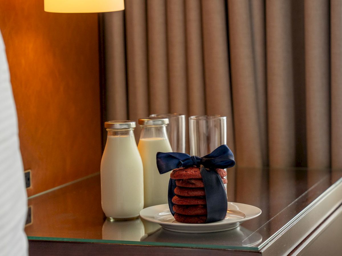 A bedside table with milk, a stack of cookies tied with a blue ribbon, under a warm lamp against closed curtains.