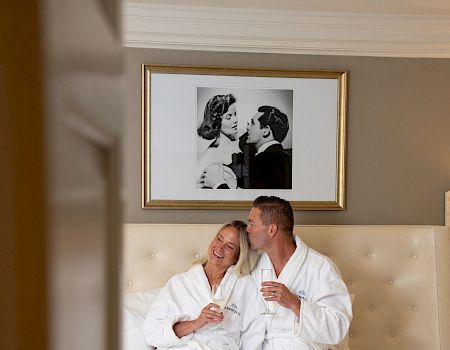 A couple in white robes is sitting on a bed, holding drinks, beneath a framed black and white photo.